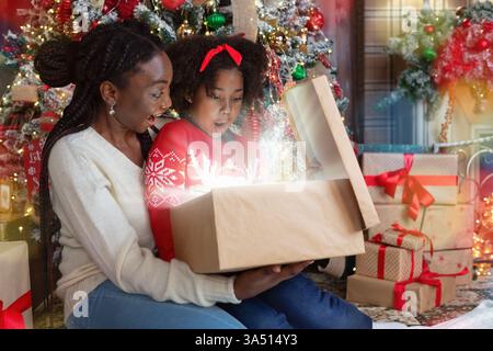Afroamerikanische Mutter und Tochter feiern Weihnachten zu Hause und öffnen eine leuchtende Geschenkbox neben einem beleuchteten Weihnachtsbaum. Ein warmer Familienmoment, der Zweisamkeit und Urlaubsfreude betont. Stockfoto