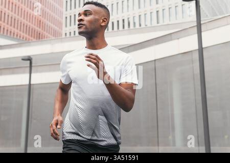 Flacher Blick auf einen schwarzen Sportler, der bei Morgenlicht in einer modernen Innenstadt läuft. Die dynamische Pose unterstreicht Sportlichkeit und Ausdauer in einer urbanen Umgebung. Ideal für Fitness-, Sportswear- und gesunde Lifestyle-Kampagnen. Stockfoto