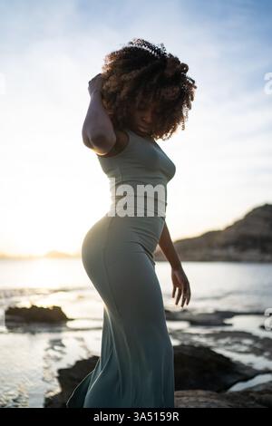 Schwarze Frau in einem Kleid mit lockigen Afro-Haaren, die bei Sonnenuntergang am Strand auf einem Felsen steht. Dramatisches Outdoor-Lifestyle-Image für Mode, Schönheit und Reisekontexte. Perfekt für Beauty- und Sommerkampagnen. Stockfoto
