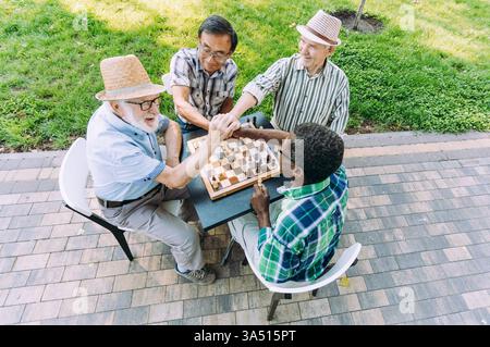 Verschiedene Seniorinnen sitzen bei Tageslicht an einem Parktisch und verbinden sich in einer Geste der Kameradschaft mit den Händen. Diese Freizeitszene im Freien ist ideal für Altersvorsorge, Freundschaft und Community Lifestyle, mit einer Atmosphäre entspannter Brettspiele und Unterhaltung im Park. Stockfoto