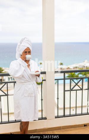 Die hispanische Frau mittleren Alters in einem weißen Bademantel mit einem Handtuch auf dem Kopf genießt Kaffee auf einem Balkon mit Meerblick. Ein entspannendes Bild des Lebensstils für Urlaubs-, Wellness- und Urlaubsangebote. Stockfoto