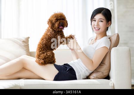 Eine junge Chinesin spielt mit einem Pudel zu Hause in einem entspannten Wohnraum. Dieser fröhliche Moment ist ideal für den Familienstil und das Leben zu Hause. Stockfoto