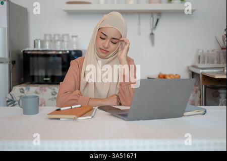 Eine gestresste asiatische Muslimbrau sitzt an einem Küchentisch und arbeitet an einem Auftrag. Die Szene spiegelt die Fernarbeit und den Druck wider, den das Studium oder die freiberufliche Tätigkeit von zu Hause aus mit sich bringt. Ein anpassbares Lifestyle-Bild mit Fokus auf Konzentration und Multitasking. Stockfoto