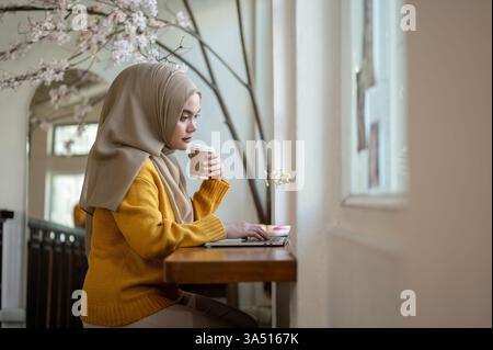 Seitenansicht einer asiatischen muslimischen Frau, die einen Hijab trägt und Kaffee trinkt, während sie einen Laptop in einem Café benutzt. Sie verbindet Arbeit und Freizeit in einem entspannten Café. Ideal für Remote-Arbeiten, freiberufliche Tätigkeiten und multikulturelle Lifestyle-Bilder. Stockfoto