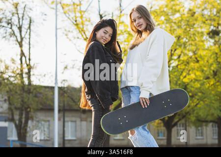 Lächelnde asiatische Frau in einer Jeansjacke berührt ihren Kopf, während sie bei Tageslicht mit einer kaukasischen Freundin in einem Park steht. Die beiden Freunde teilen einen fröhlichen, lockeren Moment. Geeignet für Freundschaft, Lifestyle und Campus- oder Outdoor-Grafiken. Stockfoto