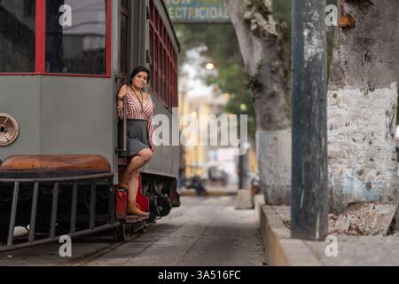 Lächelnde hispanische Frau mit dunklen kurzen Haaren in trendigem Outfit steht während ihrer Reise auf den Stufen einer alten Straßenbahn in der Stadt. Dieses reisebereite Bild vereint urbanen Stil mit sonniger, fröhlicher Stimmung. Ideal für Reisekampagnen, Straßenmode und Stadtrundfahrten. Stockfoto