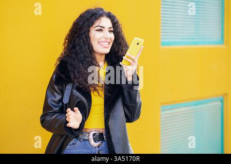 Lächelnde arabische Frau mit lockigem Haar in schwarzer Lederjacke, die Sprachnachrichten auf dem Smartphone aufnimmt und wegblickt, während sie gegen gelbes W steht Stockfoto