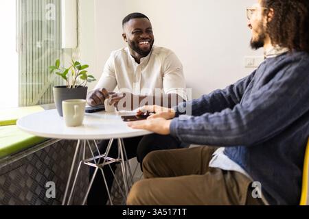 Zwei Männer unterhalten sich bei einem Kaffee in einem modernen Zuhause. Die entspannte, abwechslungsreiche Szene vermittelt Freundschaft, Wärme und alltägliche Momente. Ideal für Lifestyle, multikulturelle Werbung und soziale Interaktion. Stockfoto