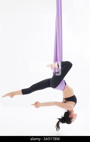 Junge Chinesin, die Luft Yoga praktiziert, in einer Hängematte in der Luft. Die athletische Pose vermittelt Balance und Vitalität, perfekt für Wellness- und Fitnesskampagnen. Stockfoto