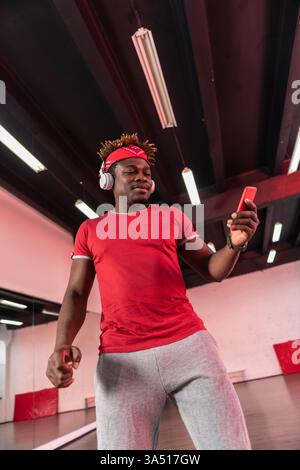 Schwarzer Mann in Kopfhörern benutzt ein Smartphone, während er in einem Tanzstudio tanzt. Der energiegeladene Moment fängt Bewegungen und Musik in einem Studio-Ambiente ein. Ideal für Tanz-, Fitness- und Lifestyle-Kampagnen. Stockfoto