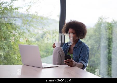 Lifestyle-Bild einer jungen Afroamerikanerin, die von zu Hause aus arbeitet. Sie sitzt an einem hellen Fenster mit einem offenen Laptop auf dem Tisch und einem weißen Becher in der Hand und lächelt auf den Bildschirm. Ideal für Remote-Arbeiten, Freiberufler und Home Office-Kampagnen. Stockfoto