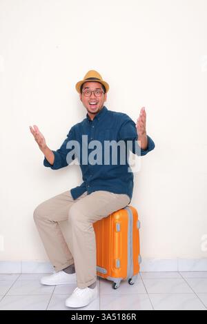 Ein asiatischer Mann sitzt mit einem Wow-Ausdruck auf seinem Koffer. Dieses Bild mit dem Thema Reisen eignet sich gut für Flughafen-, Reise- und Urlaubskontexte. Stockfoto