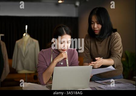 Eine fokussierte Millennials asiatische Modedesignerin sitzt mit einem Laptop an ihrem Studiotisch und arbeitet mit einem Assistenten an Designprojekten zusammen. Diese professionelle Studioszene beleuchtet kreatives Modedesign, Teamwork und unternehmerische Energie in einem modernen Boutique-Ambiente. Stockfoto