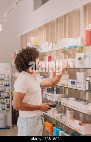 Bärtiger Mann mit Afro, der in einem Apothekengang ein Arzneimittelflaschenetikett liest. Ideal für Gesundheitsthemen und Konsumentengesundheit, mit Schwerpunkt auf Medikamenten und Einkaufen. Stockfoto