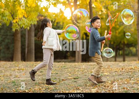 Zwei lächelnde chinesische Kinder, ein Junge und ein Mädchen, spielen mit Blasen in einem Herbstpark. Die Szene fängt unbeschwerten Outdoor-Spaß und das saisonale Laub ein. Geeignet für Familien, Aktivitäten für Kinder und Bilder des natürlichen Lebensstils. Stockfoto