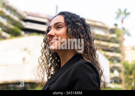 Aus einer niedrigen Seitenansicht blickt eine selbstbewusste junge hispanische Geschäftsfrau in einer schwarzen Jacke vor einem verschwommenen Stadtbild ab. Dieses zeitgenössische Porträt eignet sich hervorragend für Corporate Branding, Frauen in der Wirtschaft und städtische Karrieremöglichkeiten. Stockfoto