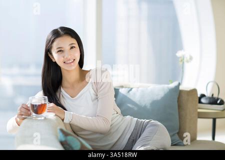 Eine fröhliche junge Chinesin genießt zu Hause eine Tasse schwarzen Tee. Der entspannte Moment findet in einem modernen Wohnzimmer statt, das sich hervorragend für Lifestyle und Wohlbefinden eignet. Perfekt für Kampagnen über Teekultur, Komfort und zeitgenössischen asiatischen Lebensstil. Stockfoto