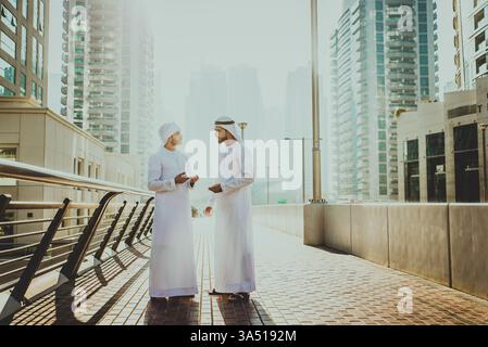 Zwei Männer aus dem Nahen Osten, die in Kanduras gekleidet sind, unterhalten sich auf einer Stadtstraße, hinter der die Skyline sichtbar ist. Freundlicher Moment, in dem geschäftliche und soziale Beziehungen in den VAE, Dubai, hervorgehoben werden. Geeignet für Besonderheiten des Lebens in der Golfstadt, Netzwerke und zeitgenössische Kultur des Nahen Ostens. Stockfoto