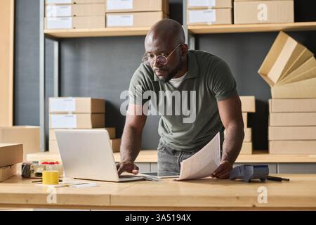 Fokussierter afrikanischer Mann in einem Lagerhaus, der Paketdaten auf einem Laptop an einem Tisch eingibt. Das Bild vermittelt Konzentration und professionelle Anstrengung in einer Logistikumgebung mit Regalen und Boxen im Hintergrund. Geeignet für Geschäfts-, Logistik- und Arbeitsplatztechnologien. Stockfoto