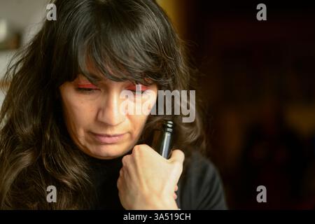 Deprimierte hispanische Frau, die zu Hause ein Glas Rotwein hält, was Stress und Einsamkeit vermittelt. Dieses stimmungsvolle Porträt kann psychische Gesundheit und emotionales Wohlbefinden in häuslichen Umgebungen veranschaulichen. Stockfoto