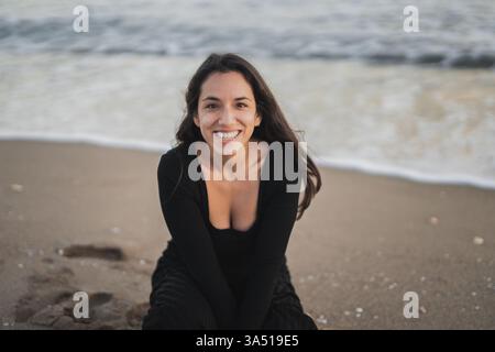 Fröhliche Mixed Race Frau mit langen dunklen Haaren in einem schwarzen Maxi-Kleid, die während des Urlaubs auf einem Sandstrand sitzt Stockfoto