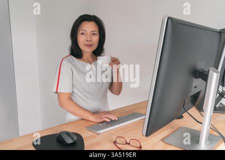 Reife asiatische Geschäftsfrau steht an einem Schreibtisch im Home Office und macht eine Pause bei einem Kaffee. Dieses professionelle Porträt eignet sich für Remote-Arbeiten, Home-Office-Setups und weibliche Führungsthemen. Eine ansprechbare Stimmung für Geschäfts- und Karrierethemen. Stockfoto