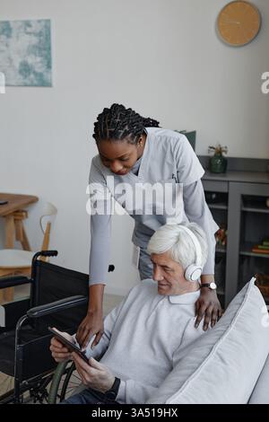 Vertikales Porträt eines weißhaarigen älteren Mannes, der zu Hause mit einer Krankenschwester eine Tablette benutzt. Diese Szene im Gesundheitswesen und betreutes Wohnen legt den Schwerpunkt auf Pflege, Zugänglichkeit und Technologie in der Altenpflege. Ideal für die Kommunikation im Gesundheitswesen, im Alter und in der häuslichen Pflege. Stockfoto