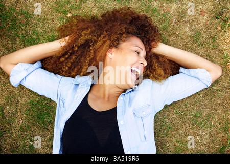 Porträt einer jungen Frau, die im Freien auf Gras lacht. Die sorglose, fröhliche Pose macht ihn ideal für Wellness, Natur und positive Lifestyle-Themen. Ideal für Redakteure, die ein brillantes, aufmunterndes Bild von Selbstvertrauen und Glück benötigen. Stockfoto