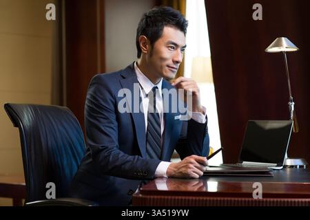 Erfolgreicher chinesischer Geschäftsmann, der tief in einer modernen Studie nachdenkt. Er sitzt mit einem Laptop an einem Schreibtisch und vermittelt Konzentration und strategische Planung. Ein professionelles Porträt, das sich für Geschäfts-, Finanz- und Führungsthemen eignet. Stockfoto