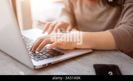 Asiatische Frau in einem Café arbeitet auf einem Laptop mit einem Smartphone auf dem Tisch. Ein freiberuflicher oder fernberuflicher Moment in einem modernen Café. Ideal für Inhalte über Coworking, Online-Produktivität und digitale Nomaden in Städten. Stockfoto