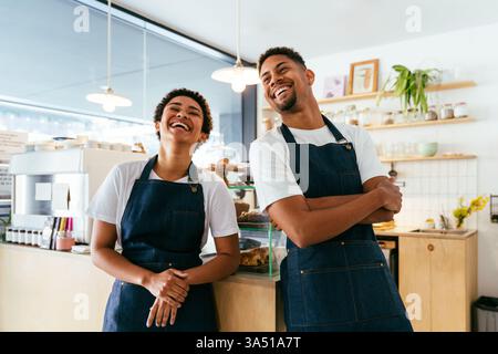 Der lächelnde hispanische Mann und die Schwarze Frau arbeiten als Barista in einem Café, die bereit sind, Gebäck und Kaffee an der Theke zu servieren. Eine vielfältige Gastfreundschaft, geeignet für Bäckerei, Cafékultur und Teamservice. Stockfoto