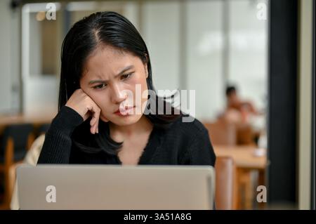 Eine verwirrte asiatische Frau sitzt mit einem Laptop, Hand am Kinn, in einem Café-Coworking-Raum. Die Szene erfasst den Fokus auf die Arbeit und den Stress, ideal für geschäftliche und freiberufliche Inhalte. Stockfoto