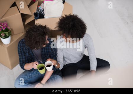 Entspanntes, multiethnisches Paar sitzt auf dem Boden in einem neuen Zuhause und genießt Kaffee zwischen ungepackten Kartons. Perfekt für Umzüge, Standortwechsel, Innenleben und Bildgestaltung. Stockfoto