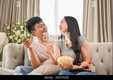 Glückliches junges asiatisches Paar genießt Popcorn, während es auf einem Sofa fernsieht. Die gemütliche Wohnzimmerszene vermittelt das häusliche Leben und die Zweisamkeit. Ideal für den privaten Lebensstil, Paare und Freizeitangebote. Stockfoto