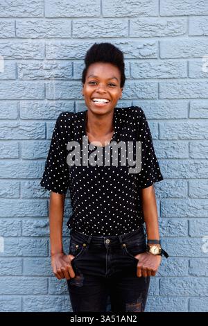 Porträt einer lächelnden Afrikanerin, die vor einer grauen Ziegelmauer steht. Lässiges, selbstbewusstes Lifestyle-Porträt, geeignet für Beauty-, Mode- und Empowerment-Botschaften. Ideal für Markeninhalte mit Fokus auf Wärme, Stil und Selbstbewusstsein. Stockfoto