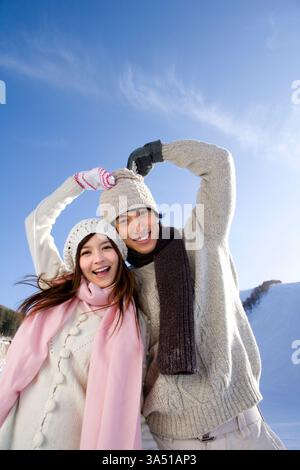 Fröhliches junges chinesisches Paar im Winter im Freien bildet mit ihren Armen eine Herzform, die Liebe und Zweisamkeit ausdrückt. Das verschneite Skigebiet, das helle Sonnenlicht und der sichtbare Kopierraum schaffen einen warmen, gemeinsamen Moment der Romantik. Stockfoto
