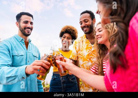 Verschiedene Freunde, die Bierflaschen auf einer sonnigen Dachterrasse anstoßen. Ein lebendiger Moment der Freundschaft, des Feierns und des städtischen gesellschaftlichen Lebens. Ideal für Lifestyle, Dating oder Reisen. Stockfoto