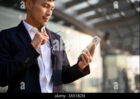 Ein erfolgreicher asiatischer Geschäftsmann der Millennials in einem formellen Anzug schaut sich sein Smartphone in einer Stadt an. Er hält seine Brille und vermittelt den Fokus und die Führungsrolle in einem digitalen Arbeitsplatz. Dieses Image eignet sich für Unternehmen, Technologie und Unternehmenskommunikation. Stockfoto