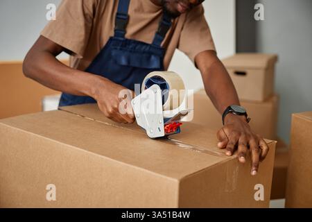 Nahaufnahme eines uniformierten afroamerikanischen Arbeiters, der Kisten packt und mit Klebeband versiegelt. Ideal für die Verlegung, das Umziehen des Büros oder die Bereitstellung von Bildern, die in Innenräumen mit Möbeln und Pappe gezeigt werden. Stockfoto