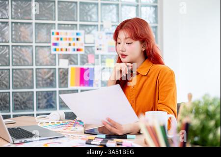 Eine fokussierte asiatische Grafikerin mit rotem Haar wählt Farbmuster an ihrem Schreibtisch in einem modernen Studio aus. Sie entwirft ein neues Projekt, das Kreativität mit durchdachter Farbauswahl verbindet. Ideal für Branding, Designprojekte und freie Illustrationskampagnen. Stockfoto