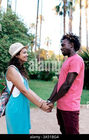 Ein junges Paar in einem Park hält Hände und teilt einen Blick, eingerahmt in einer sonnigen Umgebung im Freien. Der zärtliche Moment vermittelt Romantik und Verbindung für Lifestyle und Dating-Inhalte. Ideal für Sommer, Wochenendausflüge und Outdoor-Dating-Kampagnen. Stockfoto