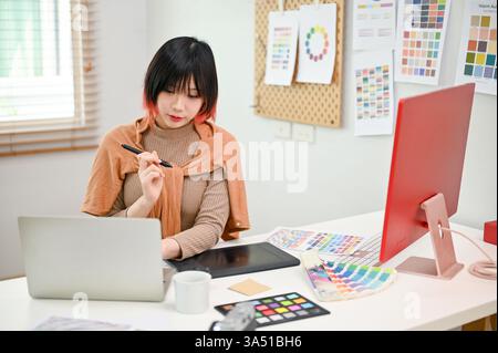 Charmante asiatische Grafikdesignerin, die in einem hellen Studio an einem Laptop arbeitet. Sie konzentriert sich auf ein neues Projekt, das Kreativität und Konzentration vermittelt. Ideal für Design-, Technologie- und freiberufliche Workflowszenen. Stockfoto