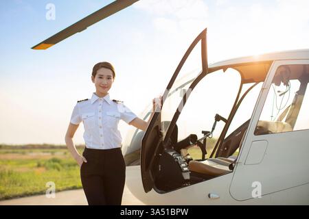 Chinesischer Hubschrauberpilot steht neben einem Hubschrauber in einer professionellen Outdoor-Umgebung. Dieses Bild zeigt Frauen in den Bereichen Luftfahrt, Luft- und Raumfahrt und MINT. Ideal für Technologie-, Transport- und Branchenkampagnen, in denen weibliche Führungsqualitäten zum Ausdruck kommen. Stockfoto