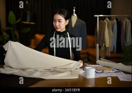 Die wunderschöne asiatische Modedesignerin der Millennials prüft in ihrem Studio den Stoff und entwickelt eine neue Kollektion. Studioszene für Modedesign, Boutiquen und kreative Brancheninhalte. Perfekt für Branding, Produkteinführungen und Designerprofile. Stockfoto