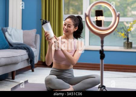 Lächelnde Chinesin nimmt zu Hause ein Video mit Ringlicht und Smartphone auf. Sie sitzt mit überkreuzten Beinen und schafft ein entspanntes Heimstudio, ideal für Vlogging und Live-Streaming. Dieses Bild passt zu Inhalten über soziale Medien, Influencer und Online-Videoerstellung. Stockfoto