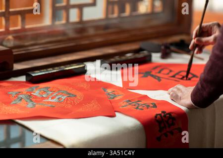 Ein hochrangiger Chinese schreibt zu Hause Kalligrafie über Couplets und hält einen festlichen Neujahrsmoment fest. Die Inneneinrichtung betont traditionelle Kultur und chinesische Schrift mit rotem Dekor. Eine Nahaufnahme, respektvolle Darstellung eines älteren Erwachsenen, der die Kunst des Schreibens genießt. Stockfoto