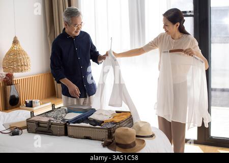 Glückliches reifes chinesisches Paar, das einen Koffer für den Urlaub in einem Hotelzimmer packt. Sie lächeln, während sie Reiseutensilien und Kleidung vorbereiten und die Zweisamkeit genießen. Diese Reise- und Lifestyle-Szene eignet sich hervorragend für Urlaubsplanung, Packen und Kampagnen, die auf Familien oder Senioren ausgerichtet sind. Stockfoto