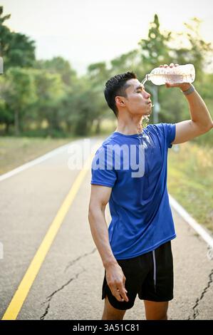 Porträt eines asiatischen Mannes in Sportbekleidung, der sich nach einem Lauf abkühlt und Wasser aus einer Flasche in einem Park auf sein Gesicht gießt. Ein erfrischender Moment, der die Feuchtigkeit und das Training im Freien unterstreicht. Stockfoto