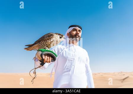 Emirati in traditioneller Kandura steht in Wüstendünen und hält einen Falken. Das eindrucksvolle Porträt zeigt die Falknerei und die Kultur des Nahen Ostens vor einer natürlichen Kulisse. Ideal für Reisen, Luxus-Lifestyle oder kulturelle Attraktionen. Stockfoto