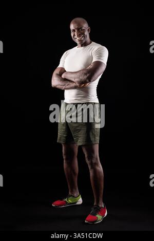 Porträt eines afroamerikanischen Sportlers, der mit überkreuzten Armen in einem Studio posiert. Der sportliche Erwachsene trägt Sportkleidung und zeigt sich der Kamera mit einer selbstbewussten Präsenz vor dunklem Hintergrund. Ideal für Fitness-, Sport- und Bodybuilding-Themen. Stockfoto